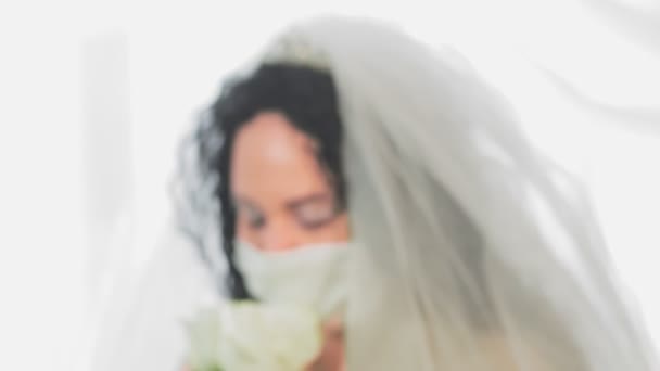 O rosto de uma noiva judia em um vestido de noiva e véu em uma máscara médica no salão da sinagoga antes da cerimônia chuppah com um buquê de flores. Do borrão à agudeza — Vídeo de Stock