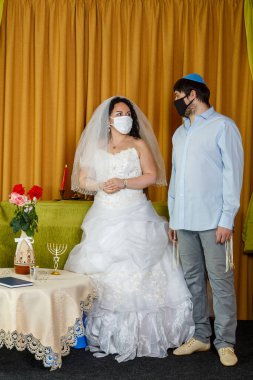 Sinagogdaki chupa töreni sırasında maskeli Yahudi gelin ve damat yan yana dururlar. Yatay fotoğraf