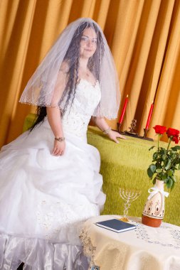 Yüzü peçeyle kaplı, bembeyaz bir elbiseyle mutlu bir Yahudi gelin, chuppah seremonisinden önce odada duruyor. Dikey fotoğraf