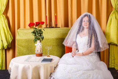 Beyaz elbiseli Yahudi bir gelin, yüzü peçeyle kaplı, chuppah seremonisinden önce çiçeklerle dolu bir masada oturuyor. Yatay fotoğraf