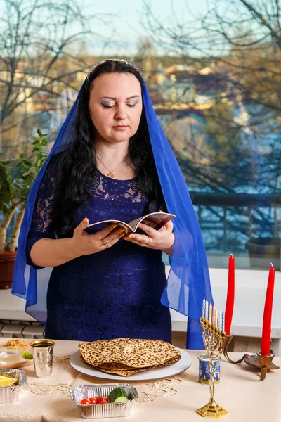 一位头戴蓝色披风的犹太妇女在逾越节的种子桌上读逾越节的 哈加德 垂直照片 — 图库照片