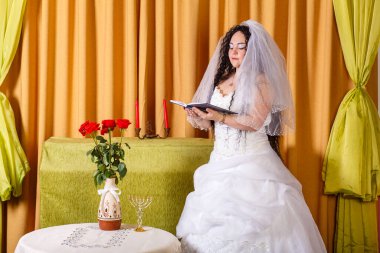 Duvağı olan beyaz gelinlikli Yahudi bir gelin salonda çiçeklerle dolu bir masada duruyor ve chuppah seremonisinden önce dua ediyor. Yatay fotoğraf