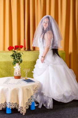 Yüzü peçeyle kaplı, bembeyaz bir elbiseyle mutlu bir Yahudi gelin, chuppah seremonisinden önce odada duruyor. Dikey fotoğraf