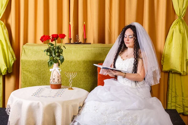 一位身穿白色婚纱婚纱的犹太新娘坐在餐桌旁 鲜花盛开 在婚礼前阅读一本祈祷书的祝福 横向照片 — 图库照片