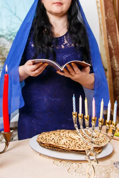 一位黑发犹太妇女，头戴蓝色披风，躺在逾越节的种子桌旁，正在阅读逾越节的《哈加德》. — 图库照片