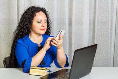 Kıvırcık esmer kadın ofiste masada oturmuş telefonla konuşuyor. Yatay fotoğraf