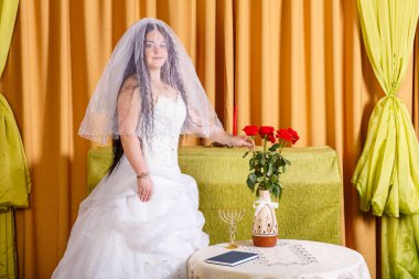 Beyaz elbiseli Yahudi bir gelin, yüzü peçeyle kaplı, çiçeklerle dolu bir masada, damadın chupa törenini beklediği bir odada duruyor. Yatay fotoğraf