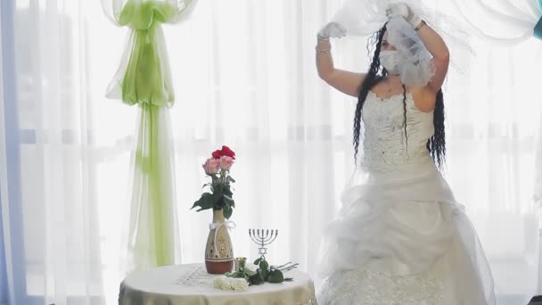 Una sposa ebrea con un abito da sposa e un velo con una maschera medica si copre la testa con un velo prima della cerimonia di Hupa. — Video Stock