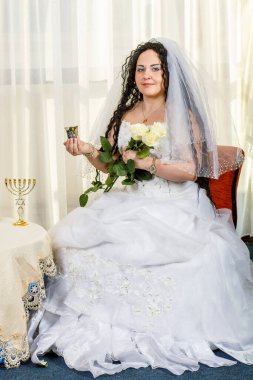 Mutlu bir Yahudi gelin, çiçeklerle dolu bir masa, bir bardak şarap ve bir buket beyaz gülle birlikte törenden önce salonda oturur. Dikey fotoğraf