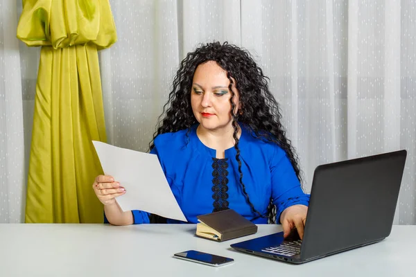 Curly brunette woman sits at the table in the office prints the text from the sheet. Horizontal photo