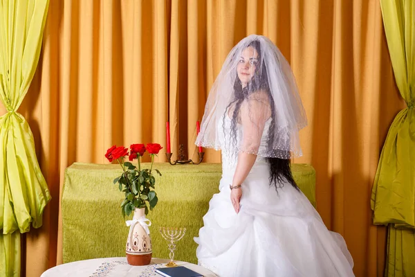 Uma noiva judia feliz em um vestido branco fofo, seu rosto coberto com um véu, fica na sala na mesa antes da cerimônia do chupa. — Fotografia de Stock
