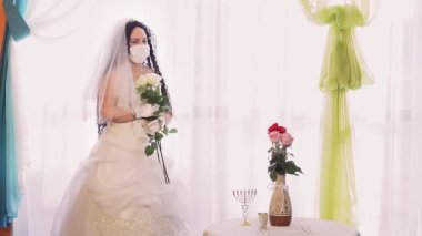 Gelinlik giymiş bir Yahudi gelin ve tıbbi maske takmış bir duvak ile sinagog salonunda bir buket beyaz gülle yüzünü çeviriyor..