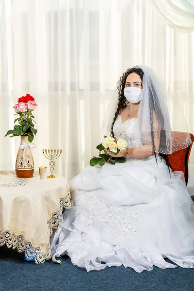 Una novia judía se sienta en una sinagoga antes de una ceremonia de chuppa durante una pandemia con una máscara médica y un ramo de flores espera al novio. —  Fotos de Stock