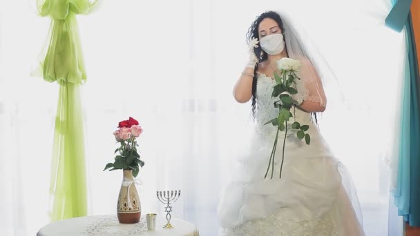 Una sposa ebrea in abito da sposa e velo che indossa una maschera medica in una sala della sinagoga prima della cerimonia della chupah parla al telefono — Video Stock