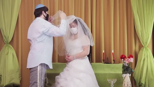 During the chuppah ceremony, a Jewish bride and groom wearing protective masks in a synagogue covers the bride with a veil — Stock Video