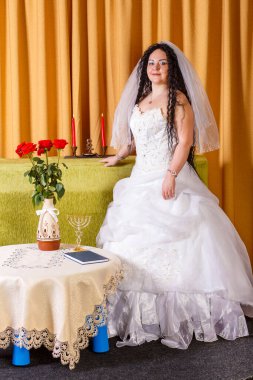 Duvağı olan beyaz gelinlikli Yahudi bir gelin Chupa seremonisinden önce çiçeklerle dolu bir masada duruyor..