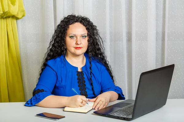 Krullend brunette vrouw zitten aan een tafel in het kantoor werken met een laptop. — Stockfoto
