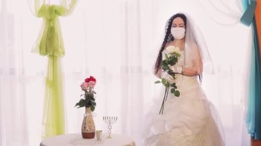 Gelinlik giymiş bir Yahudi gelin ve tıbbi maske takmış bir duvak ile sinagog salonunda bir buket beyaz gülle yüzünü çeviriyor..