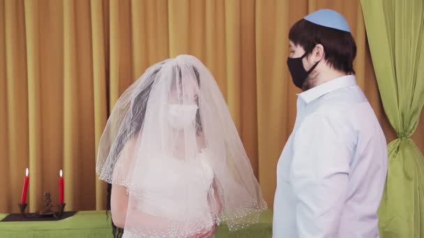 During the chuppah ceremony, a Jewish bride and groom wearing protective masks in a synagogue lifts the veil from the brides face — Stock Video
