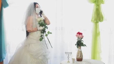 Gelinlik ve duvak takmış Yahudi bir gelin. Sinagogda tıbbi maske takıyor. Chupah töreninden önce telefonda konuşuyor.