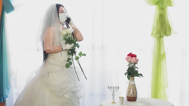 En judisk brud i bröllopsklänning och slöja klädd i en medicinsk mask i en synagogahall innan chupahceremonin talar i telefon — Stockvideo