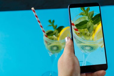 Mavi arka plandaki bir bardakta naneli salatalık ve limonlu kokteyl. Kadın eli fotoğraf çekiyor. Yatay fotoğraf