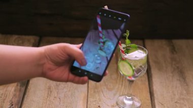 Naneli kokteyl ve cam bir salatalık ve limonlu kadın eli akıllı telefondan fotoğraf çeker.