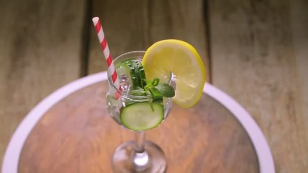 Un cocktail avec menthe concombre et citron dans un verre sur une tige sur une table en bois tourne en cercle. — Video