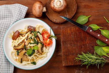 Udon, mantarlı ve ıspanaklı gri bir peçetenin üzerinde, tahtada bıçak, mantar ve yeşilliklerin yanında. Yatay fotoğraf