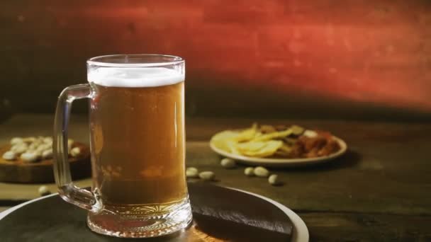 Schäumend kaltes, leichtes Bier in einem Glasbecher auf einem Holztisch mit einem Imbiss dreht sich im Kreis. — Stockvideo