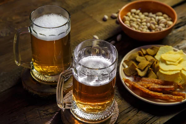 Two mugs with foamy beer and pistachios with chips on a wooden table. Horizontal photo