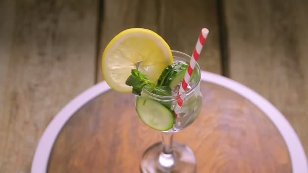 Een cocktail met komkommer munt en citroen in een glas op een steel op een houten tafel draait in een cirkel — Stockvideo