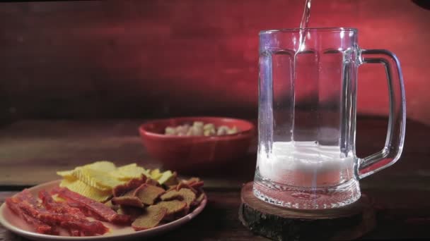 在开心果和薯片旁边的桌子上的杯子里倒出一杯泡沫的淡淡的啤酒 — 图库视频影像