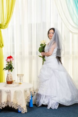 Bir Yahudi gelin, elinde bir buket beyaz gül ve çiçeklerle bir masada chuppa töreninin önünde duruyor. Dikey fotoğraf