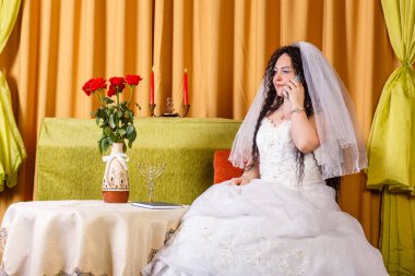 A Jewish bride in a white wedding dress with a veil sits at a table with flowers and speaks on the phone before the chuppah ceremony.