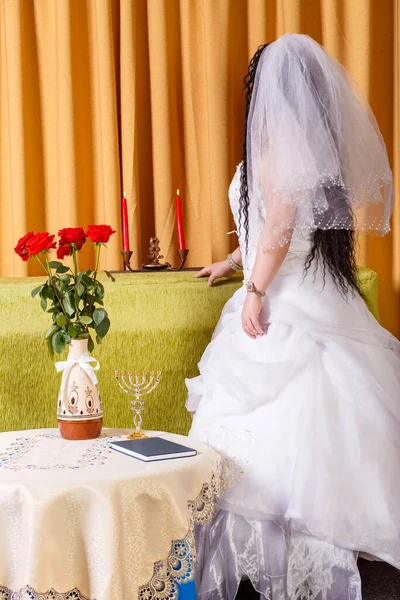 Uma noiva judia em um vestido de casamento branco com um véu fica no salão em uma mesa com flores meio viradas antes da cerimônia do chuppah.. — Fotografia de Stock