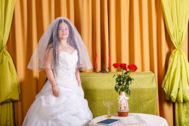 Beyaz elbiseli Yahudi bir gelin, yüzü peçeyle kaplı, çiçeklerle dolu bir masada, damadın chupa törenini beklediği bir odada duruyor. Yatay fotoğraf