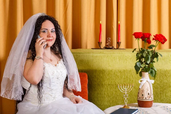 一位身穿白色婚纱婚纱的犹太新娘坐在鲜花满布的桌旁 在婚礼前通过电话交谈 横向照片 — 图库照片