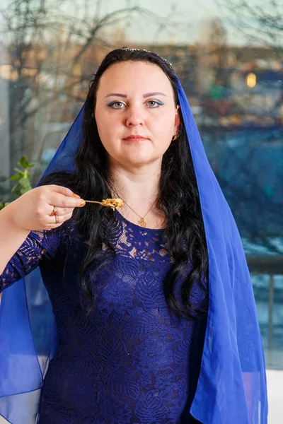 A Jewish woman with her head covered in a blue cape at the Passover Seder table eats haroset. Vertical photo