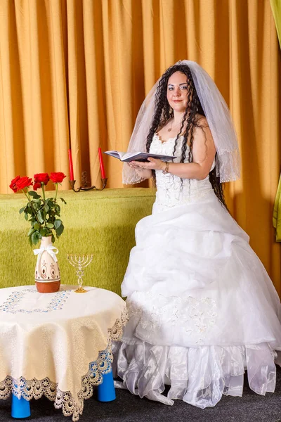 Uma noiva judia em um vestido branco, rosto velado em uma mesa com flores, lê Tegilim antes da cerimônia chupa. — Fotografia de Stock