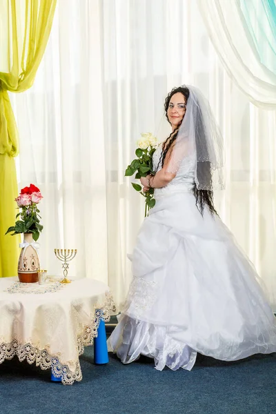 Μια ευτυχισμένη Εβραία νύφη στέκεται στην αίθουσα πριν από την τελετή Τσάπα σε ένα τραπέζι με λουλούδια με ένα μπουκέτο λευκά τριαντάφυλλα στα χέρια της. — Φωτογραφία Αρχείου