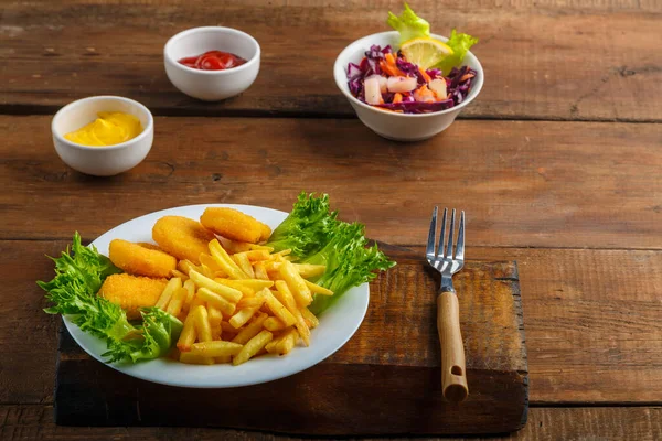 French fries with chicken nuggets next to cheese sauce and ketchup in a gravy boat and salad on a wooden table next to a fork. Horizontal photo