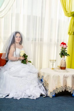 Yüzü beyaz güllerle kaplı mutlu bir Yahudi gelin, çiçeklerle dolu bir masada Huppa ayini yapmadan önce sinagogda oturur. Dikey fotoğraf