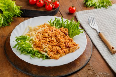 Gri bir peçete, çatal ve vişneli domateslerin yanında, ahşap bir masada yeşillerle süslenmiş bir tabak bolonez makarnası. Yatay fotoğraf
