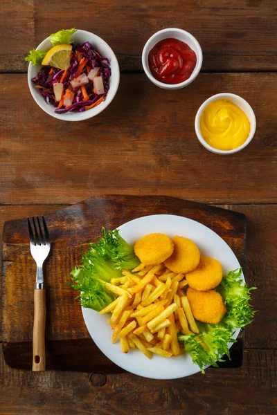 French fries with chicken nuggets near cheese sauce and ketchup in a gravy boat on a wooden table. Vertical photo