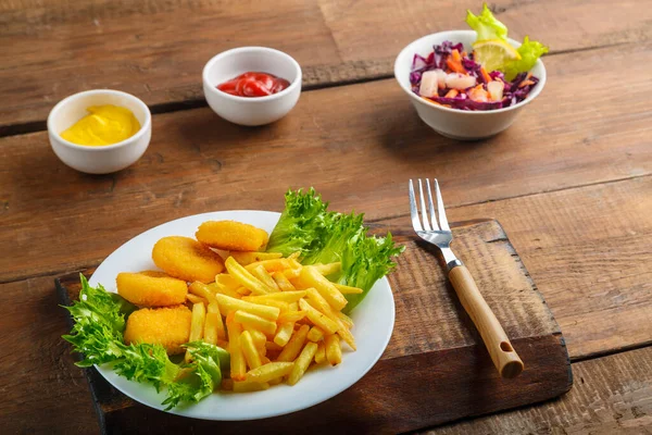 French fries with chicken nuggets next to cheese sauce and ketchup in a gravy boat and salad on wooden planks next to a fork. Horizontal photo