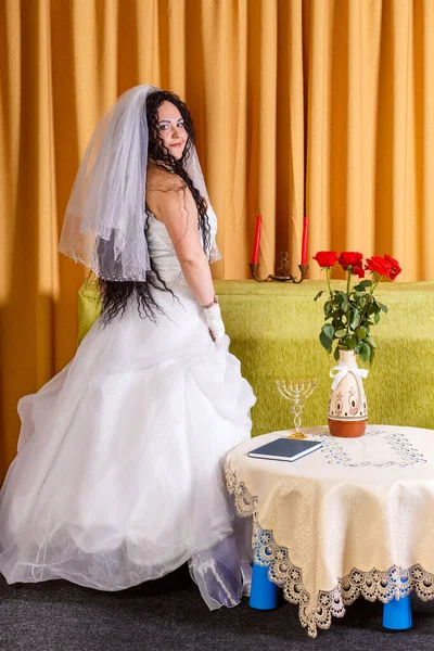 Seorang pengantin Yahudi dengan gaun pengantin putih dengan kerudung berdiri di aula di atas meja dengan bunga setengah berbalik sebelum upacara chuppah. — Stok Foto
