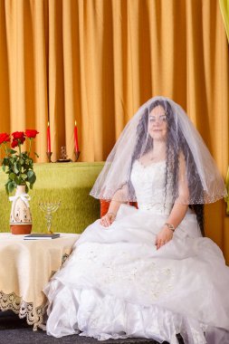 Beyaz elbiseli Yahudi bir gelin, yüzü peçeyle kaplı, chuppah seremonisinden önce çiçeklerle dolu bir masada oturuyor. Dikey fotoğraf