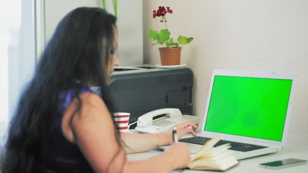 Una mujer morena trabaja en una computadora y escribe un diario en su oficina en casa con su espalda a la cámara de pantalla verde — Vídeos de Stock