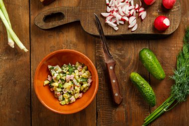 Okroshka kil bir tabakta, tahta bir masada, tahtalarda sebzelerin ve bıçağın yanında. Yatay fotoğraf